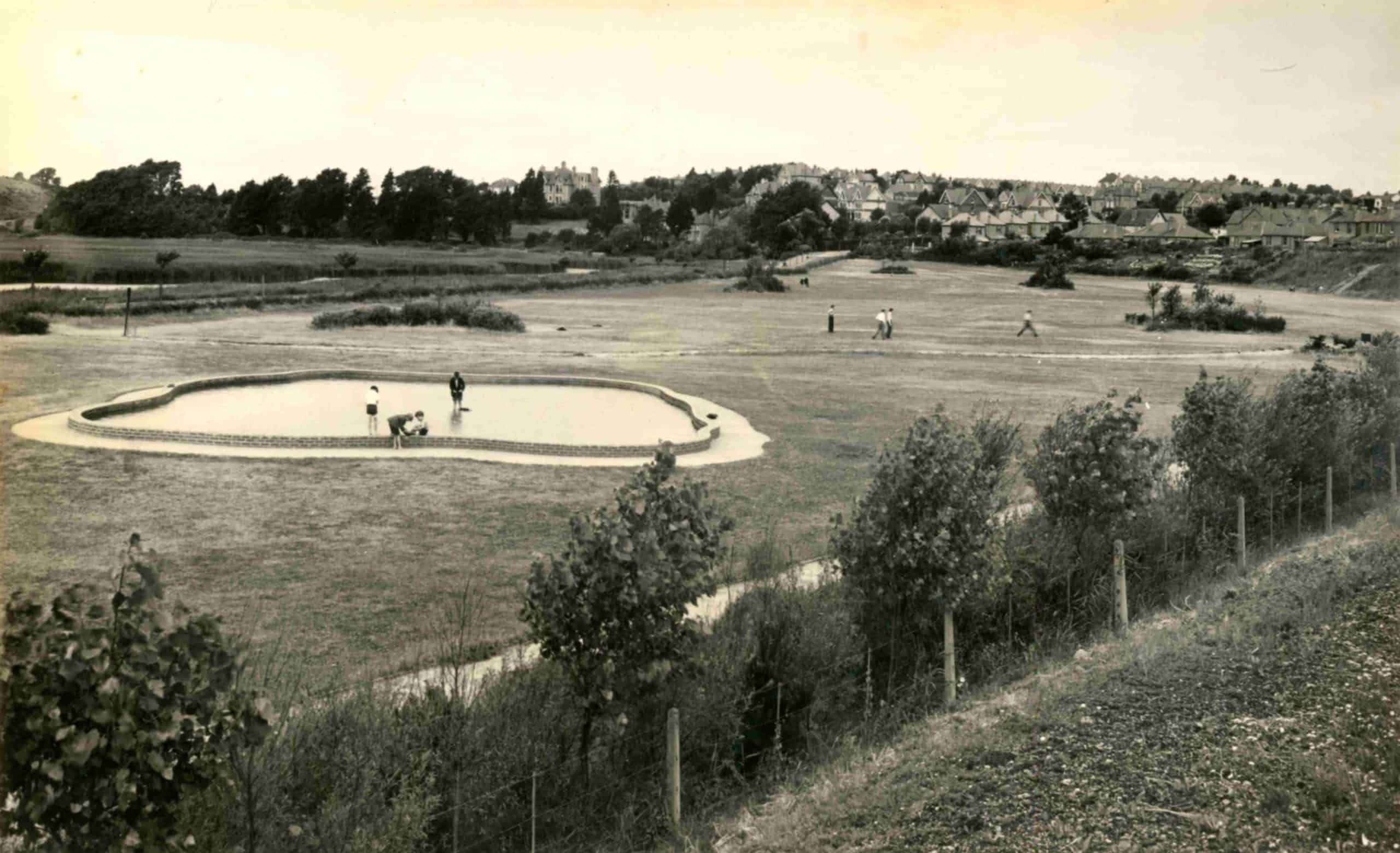 Vintage black and white photo of a park with people scattered around a pond, surrounded by open grassy areas and a distant housing development.