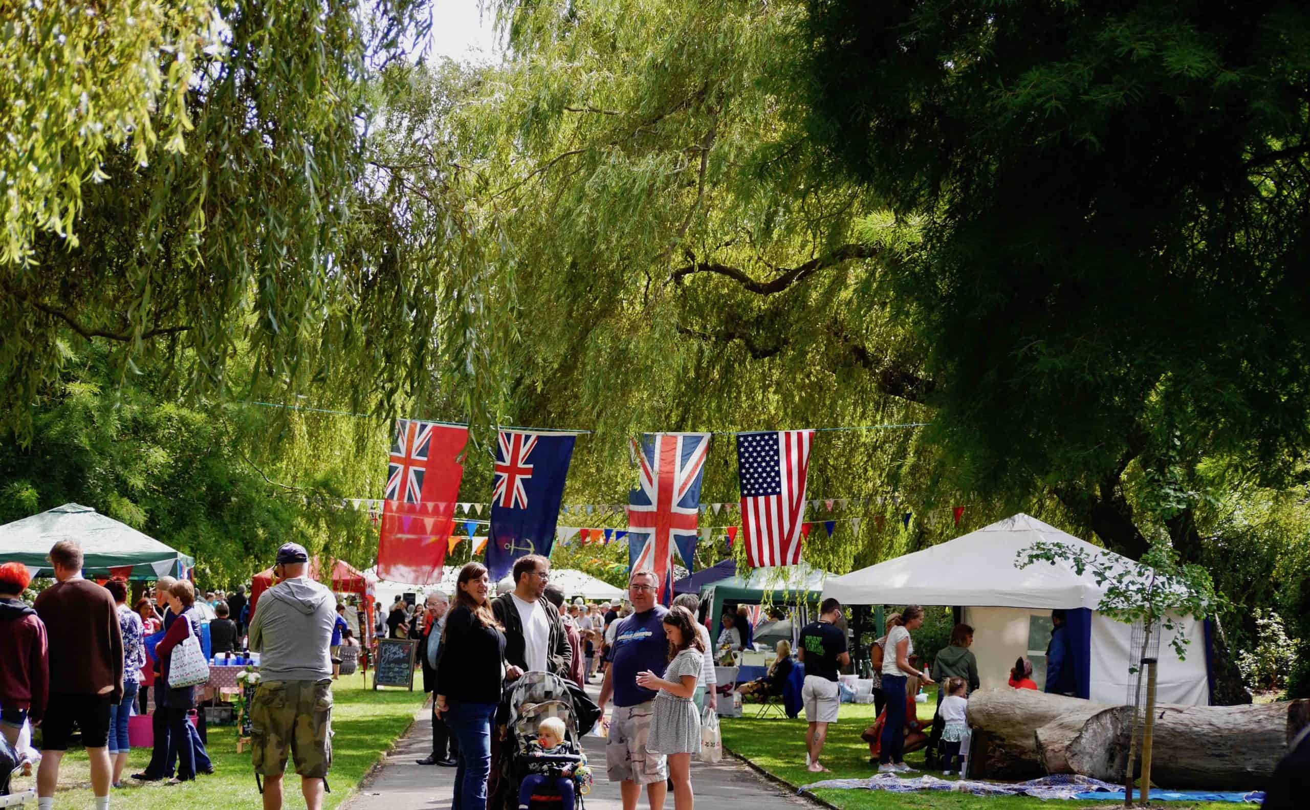 People mingle at a lively outdoor event in a park adorned with British and American flags, true to the WWII style. Stalls, trees, and a sunny sky enhance the festive atmosphere.