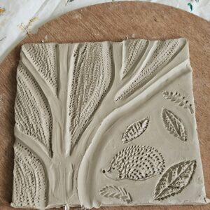 A square clay slab features a raised relief design depicting a hedgehog surrounded by various textured leaves and floral patterns, all carved into the wet clay surface.